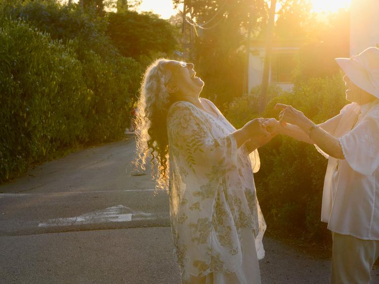 Zwei Frauen halten sich an den Händen und lachen, im Hintergrund die untergehende Sonne.