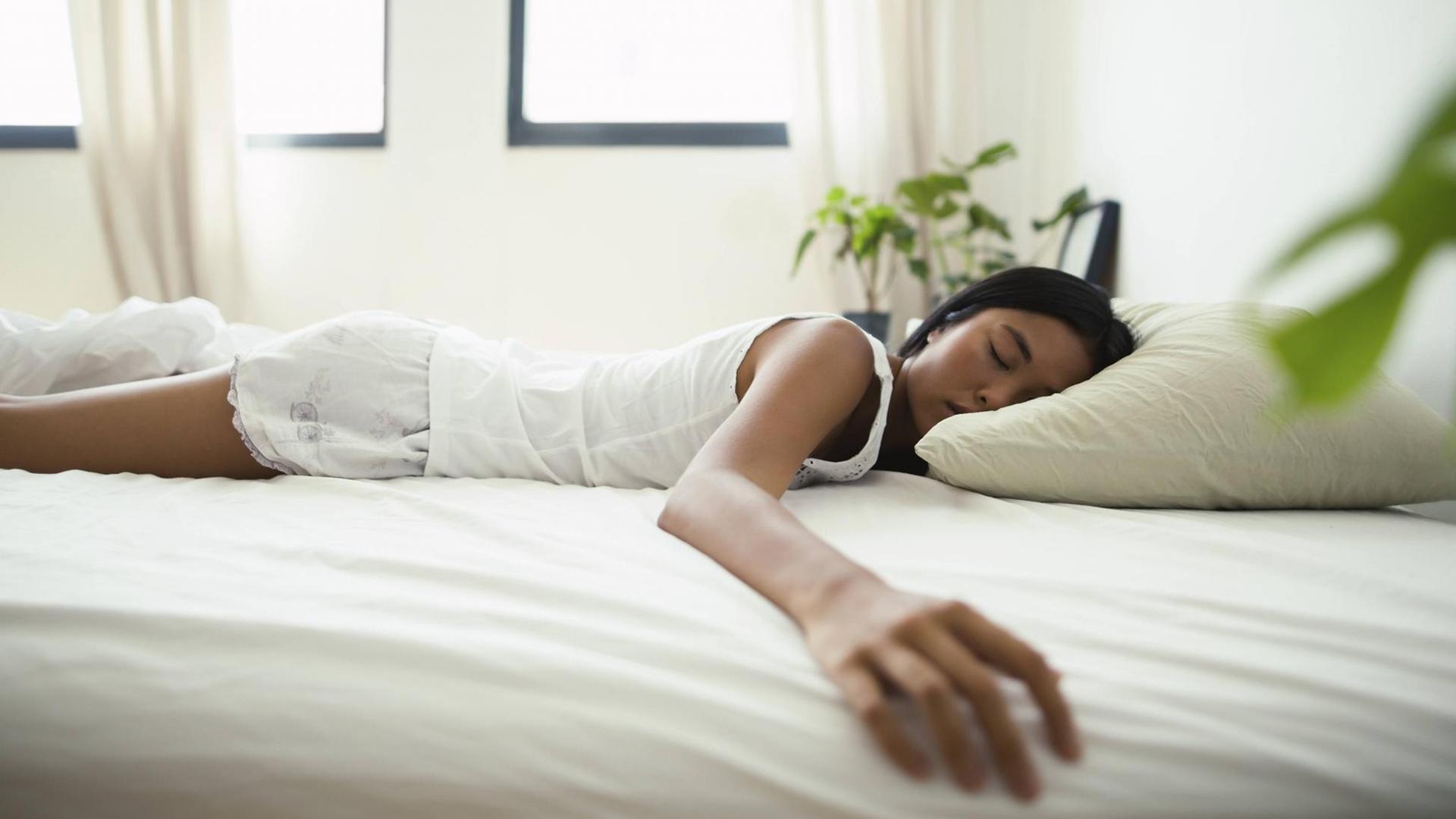 Eine Frau liegt in einem Bett und schläft. Sie liegt dabei auf dem Bauch und streckt einen Arm zur Seite aus. Eine Frau liegt in einem Bett und schläft. Sie liegt dabei auf dem Bauch und streckt einen Arm zur Seite aus.