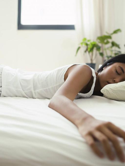 Eine Frau liegt in einem Bett und schläft. Sie liegt dabei auf dem Bauch und streckt einen Arm zur Seite aus.