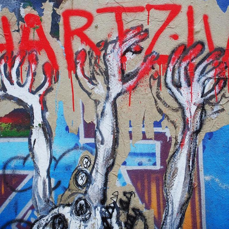 Ein unbekannter Street-Art-Künstler hat auf einer Mauer an einem besetzten Haus in Berlin im Bezirk Mitte dieses Bild geschaffen, das seine Meinung zu den Hartz IV Gesetzen drastisch wiedergibt: Verzwifelte HÄnde greifen nach dem roten Schriftzug Hartz IV, im Hintergrund sind Grabkreuze zu sehen.