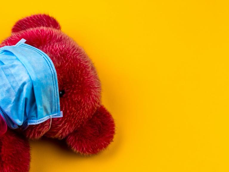 Ein roter Teddybär mit Mundschutz auf gelben Untergrund.