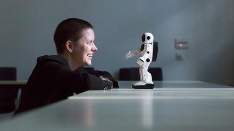 Die Philosophin Janina Loh sitzt am Tisch und blickt auf einen kleinen weissen Roboter ihr gegenüber.