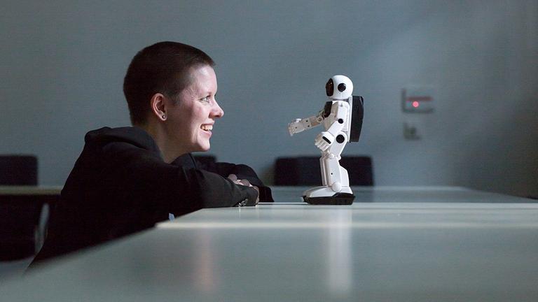 Die Philosophin Janina Loh sitzt am Tisch und blickt auf einen kleinen weissen Roboter ihr gegenüber. Die Philosophin Janina Loh sitzt am Tisch und blickt auf einen kleinen weissen Roboter ihr gegenüber.