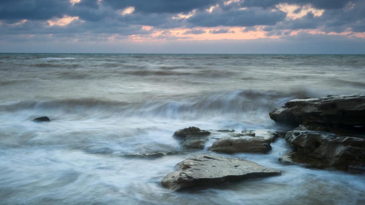 Das bewegte Meer vom Strand aus fotografiert unter wolkigem Himmel mit Abendröte. Im Vordergrund schwarzer Fels. Das bewegte Meer vom Strand aus fotografiert unter wolkigem Himmel mit Abendröte. Im Vordergrund schwarzer Fels.