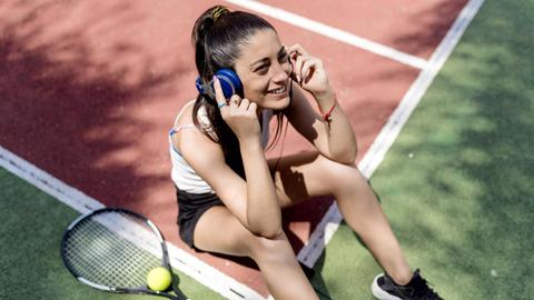 Eine lächelnde Frau sitzt in Sportkleidung auf dem Tennisplatz und hört Musik über Kopfhörer, während ihr Schläger neben ihr liegt.