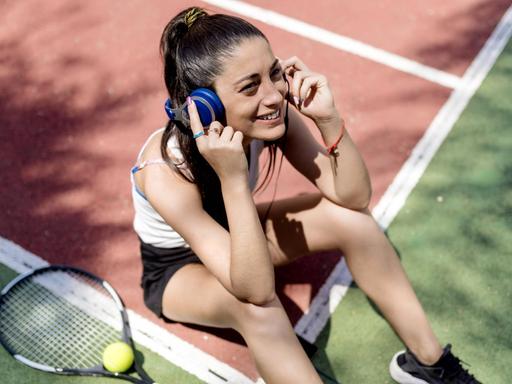 Eine lächelnde Frau sitzt in Sportkleidung auf dem Tennisplatz und hört Musik über Kopfhörer, während ihr Schläger neben ihr liegt.