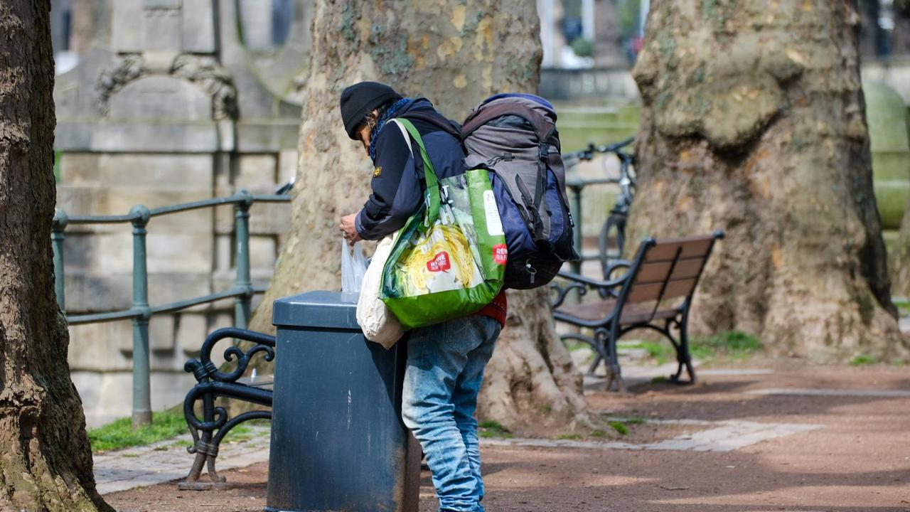 Ein Mann mit Turnschuhen und schlecht sitzenden Jeans durchsucht einen Abfalleimer an einer Parkbank. Er trägt mehrere große Plastiktaschen und einen Rucksack über der Schulter. Ein Mann mit Turnschuhen und schlecht sitzenden Jeans durchsucht einen Abfalleimer an einer Parkbank. Er trägt mehrere große Plastiktaschen und einen Rucksack über der Schulter.