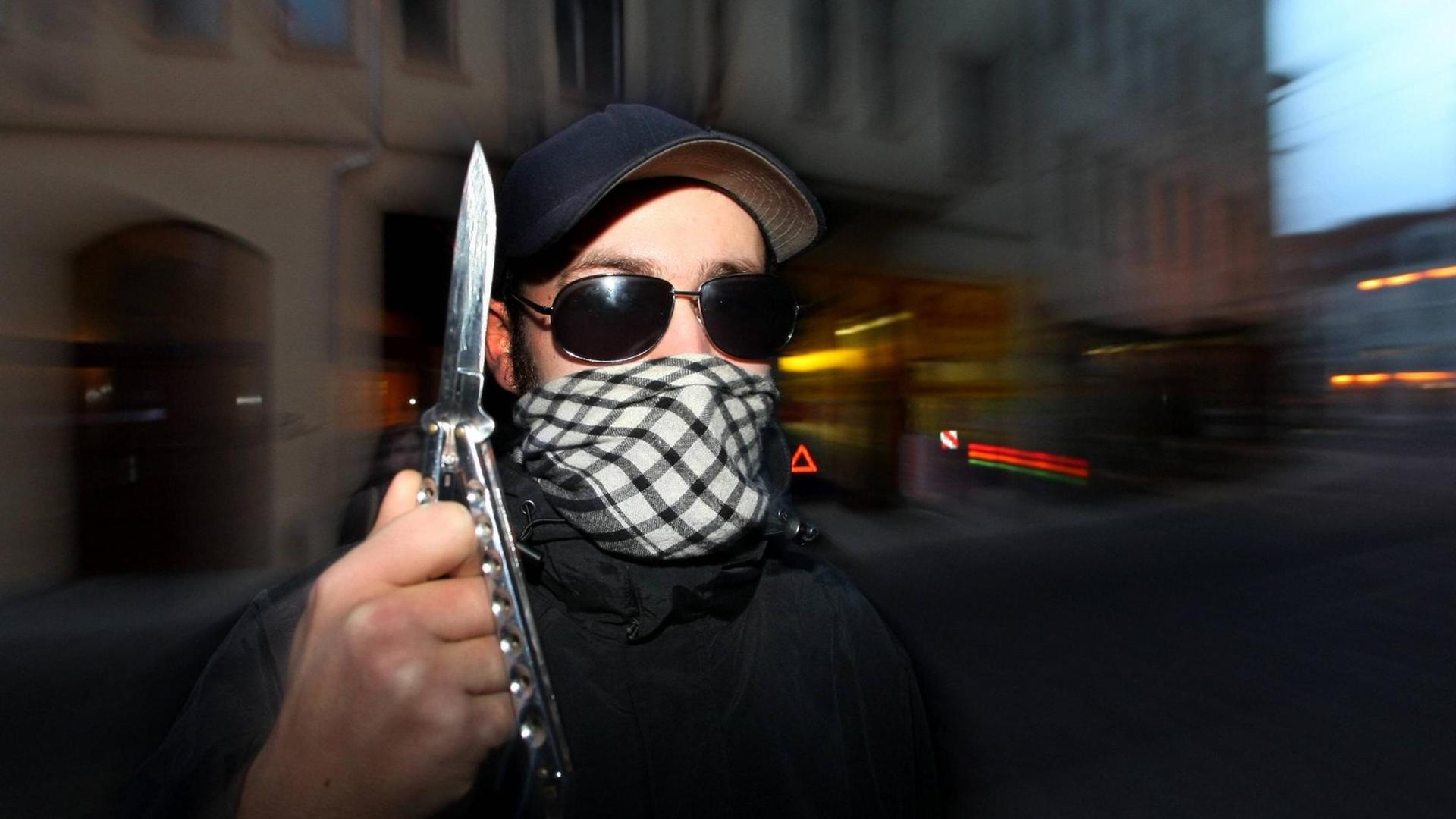Vermummter Jugendlicher mit Schirmmütze und Sonnenbrille hält ein Butterflymesser in die Höhe. Vermummter Jugendlicher mit Schirmmütze und Sonnenbrille hält ein Butterflymesser in die Höhe.