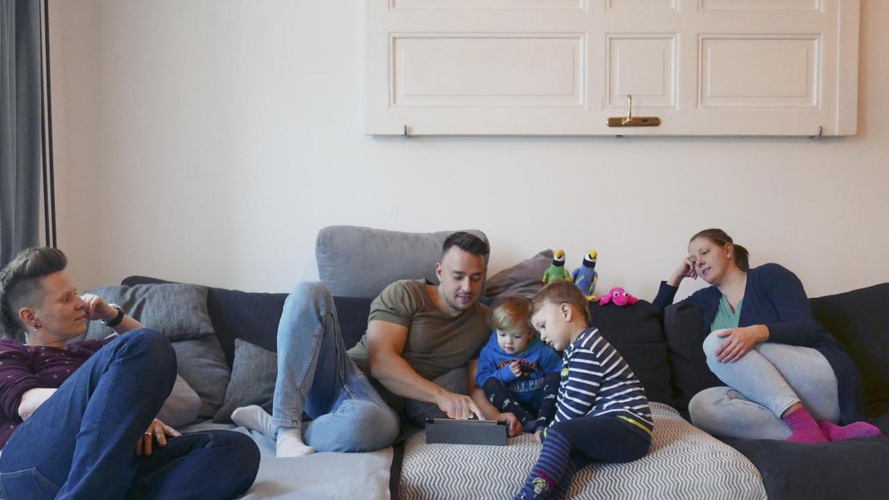 Eine Familie, bestehend aus zwei Müttern, einem Vater und zwei Kindern sitzen zusammen auf der Couch. Die Kinder schauen mit dem Vater auf ein Tablet. Eine Familie, bestehend aus zwei Müttern, einem Vater und zwei Kindern sitzen zusammen auf der Couch. Die Kinder schauen mit dem Vater auf ein Tablet.