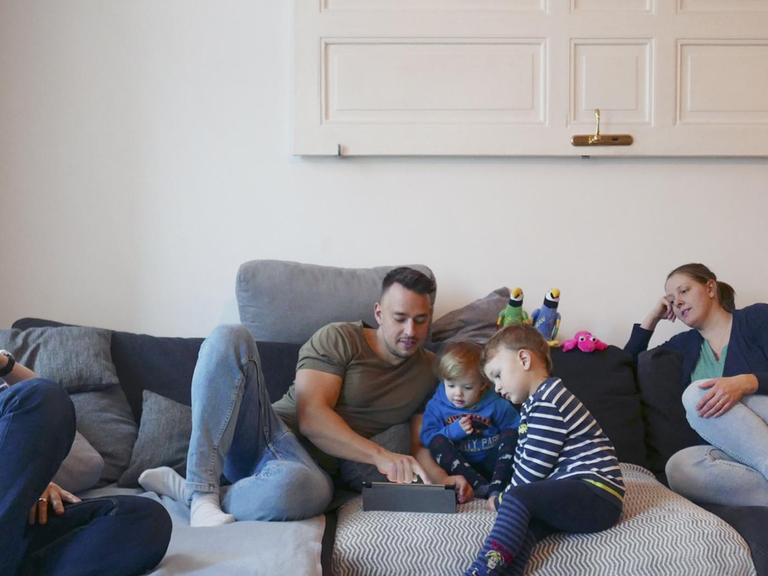 Eine Familie, bestehend aus zwei Müttern, einem Vater und zwei Kindern sitzen zusammen auf der Couch. Die Kinder schauen mit dem Vater auf ein Tablet.