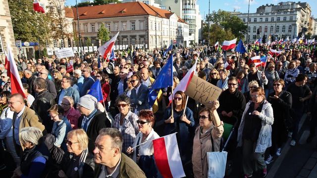 Eine große Menschenmenge mit Transparenten und polnischen Fahnen zieht eine Straße entlang. Dahinter sieht man mehrere Gebäude.