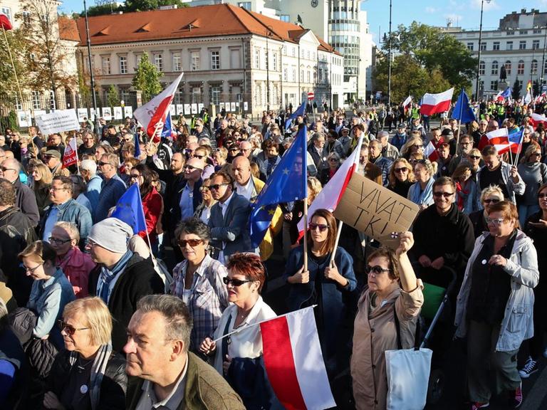 Eine große Menschenmenge mit Transparenten und polnischen Fahnen zieht eine Straße entlang. Dahinter sieht man mehrere Gebäude.