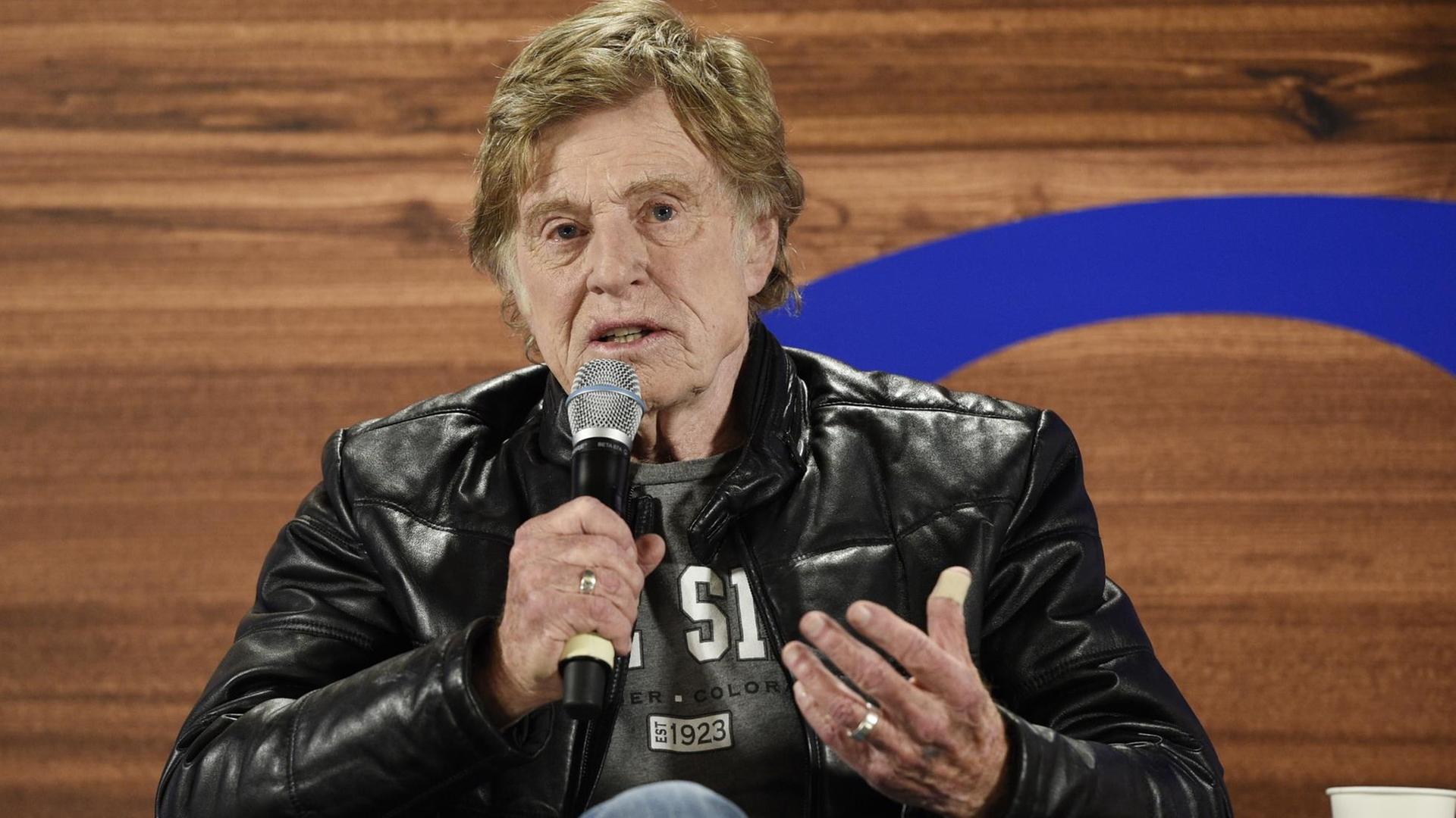 Redford sitzt in einem Stuhl in einer schwarzen Lederjacke vor einer holzgetäfelten Wand und spricht in ein Mikrofon. Redford sitzt in einem Stuhl in einer schwarzen Lederjacke vor einer holzgetäfelten Wand und spricht in ein Mikrofon.