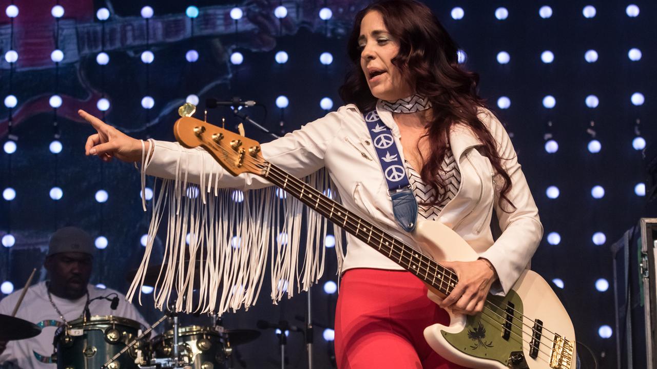 Eine Frau mit weißer Fransenjacke und Bass steht auf einer Bühne.
