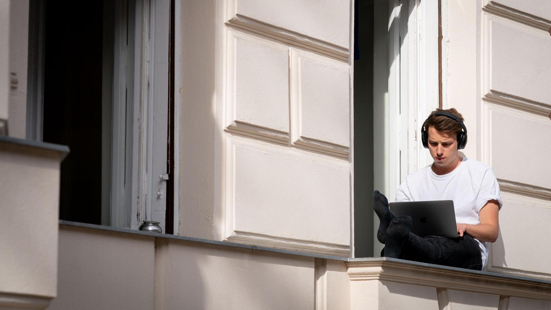 Ein Mann sitzt in Berlin auf einer Fensterbank in der Sonne und arbeitet an seinem Laptop.