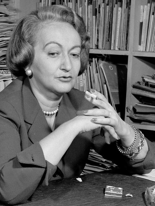 Alba de Céspedes an einem Schreibtisch, mit Zigarette in der Hand vor einem Regal mit vielen Zeitungen