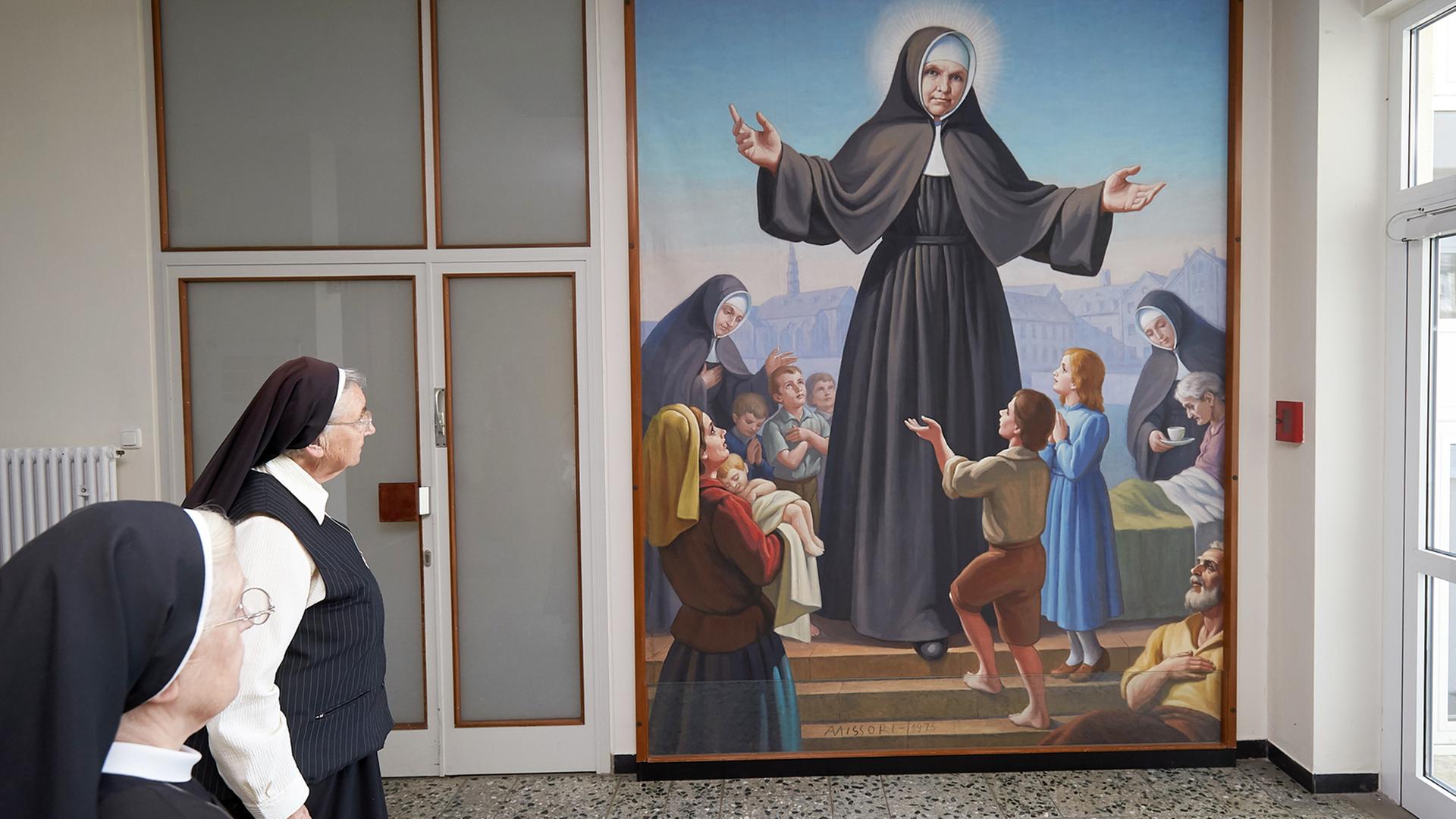 Zwei Ordensschwestern stehen vor einem Gemälde, auf dem ist Katharina Kaser zu sehen mit ausgebreiteten Armen und Heiligenschein