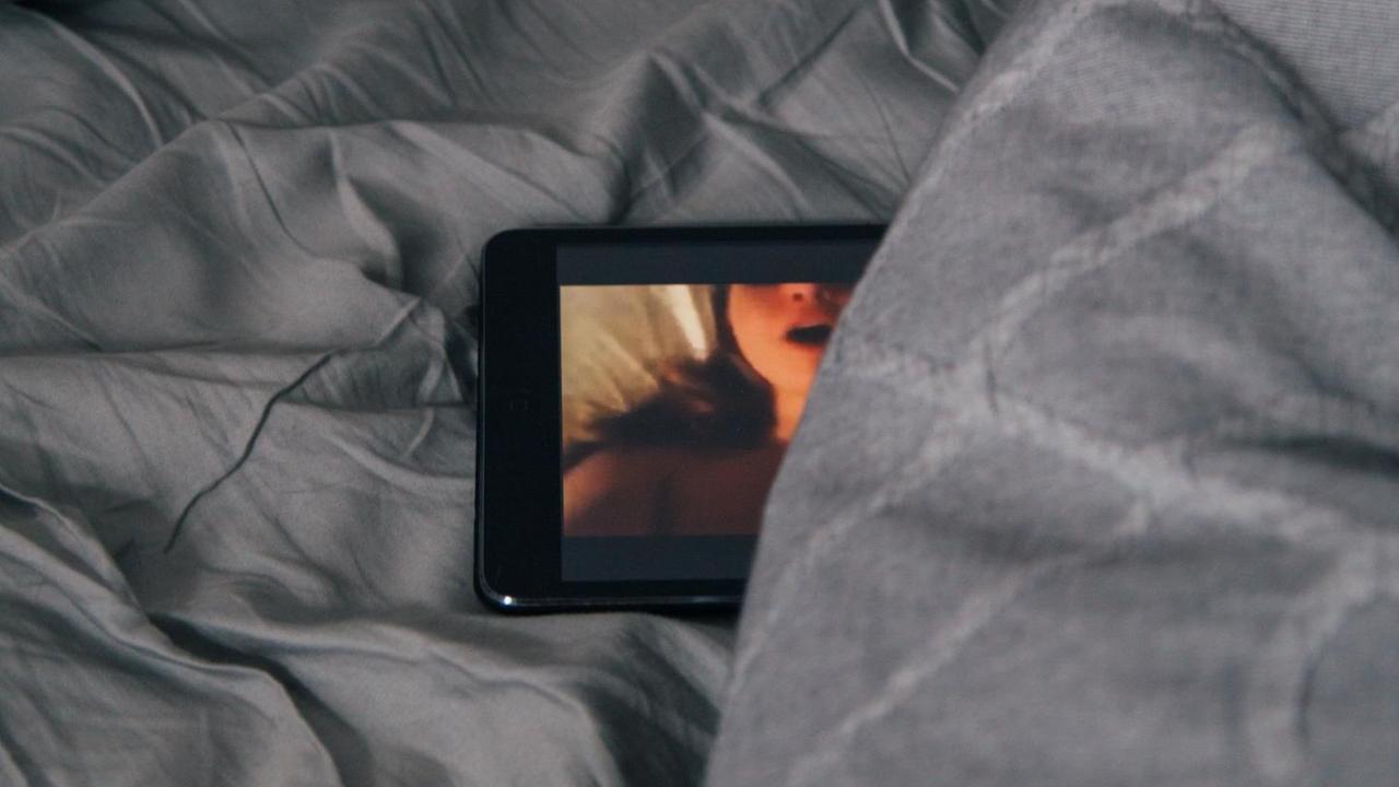 Ein Smartphone-Bildschirm schaut teilweise unter einer Bettdecke hervor. Auf dem Bildschirm ist eine stöhnende Frau beim Sex zu sehen. Ein Smartphone-Bildschirm schaut teilweise unter einer Bettdecke hervor. Auf dem Bildschirm ist eine stöhnende Frau beim Sex zu sehen.