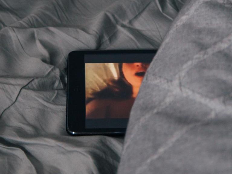 Ein Smartphone-Bildschirm schaut teilweise unter einer Bettdecke hervor. Auf dem Bildschirm ist eine stöhnende Frau beim Sex zu sehen.