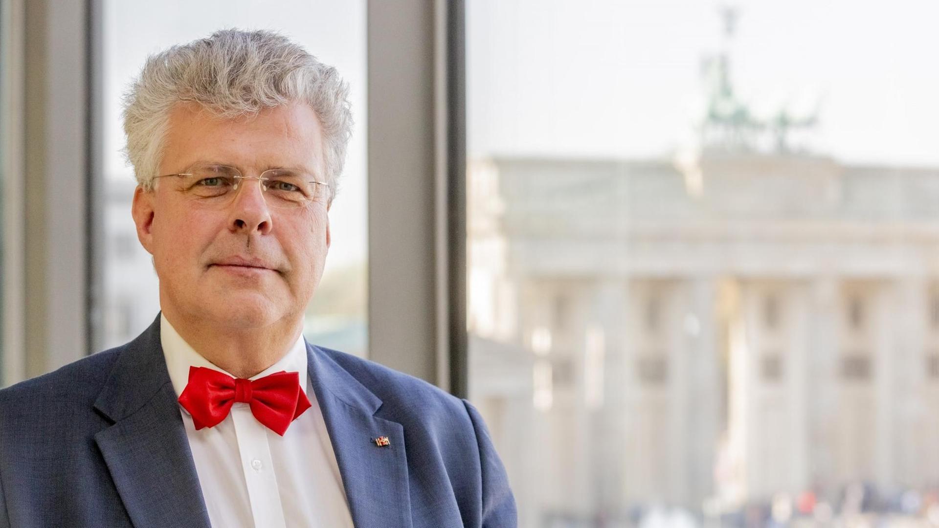 Porträtaufnahme von Christian Höppner. Er trägt eine rote Fliege, ein weißes Hemd und ein blaues Sakko. Hinter ihm ist eine Fensterfront zu erkennen, durch die man das Brandenburger Tor in Berlin sieht. Porträtaufnahme von Christian Höppner. Er trägt eine rote Fliege, ein weißes Hemd und ein blaues Sakko. Hinter ihm ist eine Fensterfront zu erkennen, durch die man das Brandenburger Tor in Berlin sieht.
