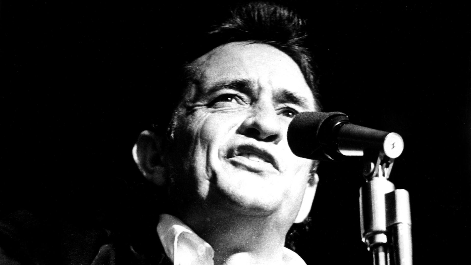 Die Schwarzweiß-Aufnahme zeigt den Sänger Johnny Cash, wie er in ein Mikrofon singt. Die Schwarzweiß-Aufnahme zeigt den Sänger Johnny Cash, wie er in ein Mikrofon singt.