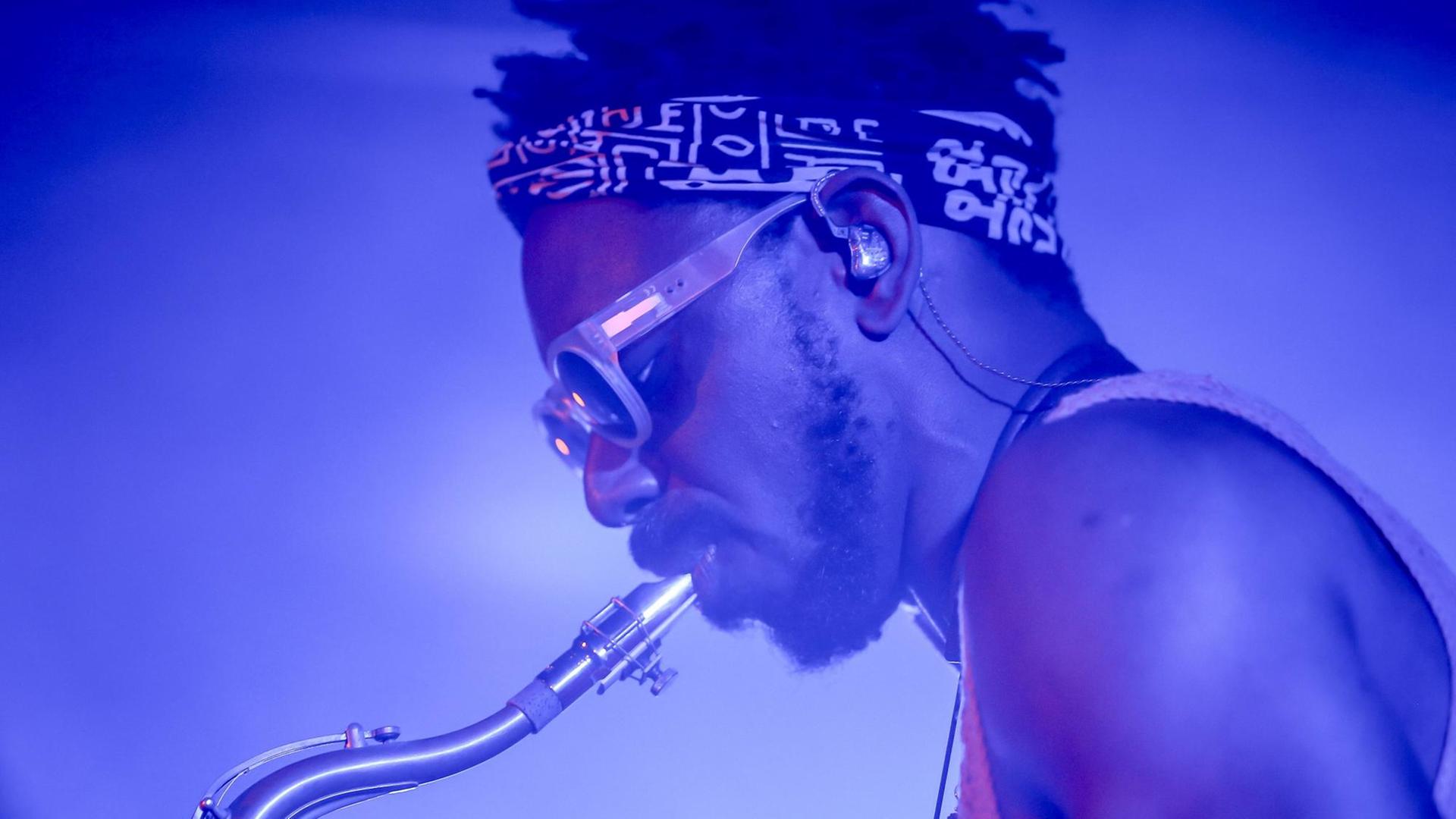 Ein Mann mit Sonnenbrille und Stirnband mit afrikanisch anmutender Musterung spielt Tenorsaxofon. Er ist in blaues Licht gehüllt und im Profil zu sehen. Ein Mann mit Sonnenbrille und Stirnband mit afrikanisch anmutender Musterung spielt Tenorsaxofon. Er ist in blaues Licht gehüllt und im Profil zu sehen.
