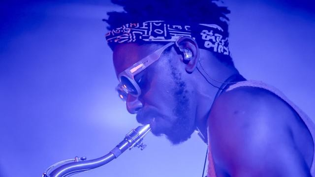 Ein Mann mit Sonnenbrille und Stirnband mit afrikanisch anmutender Musterung spielt Tenorsaxofon. Er ist in blaues Licht gehüllt und im Profil zu sehen.