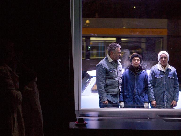 Drei Menschen stehen vor einem Schaufenster und schauen in den Raum hinein. Hinter dem Fester befindet sich ein Mensch im Dunkeln und schaut hinaus.
