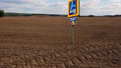 Ein Schild mit der Aufschrift "Aldi Süd" steht auf einem Acker und soll Landgrabbing, den Aufkauf von Ackerfläche im großen Stil durch Konzerne, symbolisieren.