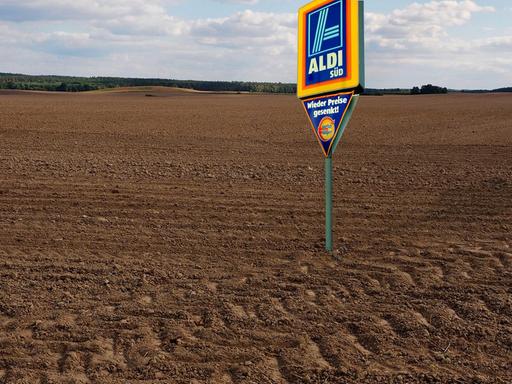 Ein Schild mit der Aufschrift "Aldi Süd" steht auf einem Acker und soll Landgrabbing, den Aufkauf von Ackerfläche im großen Stil durch Konzerne, symbolisieren.