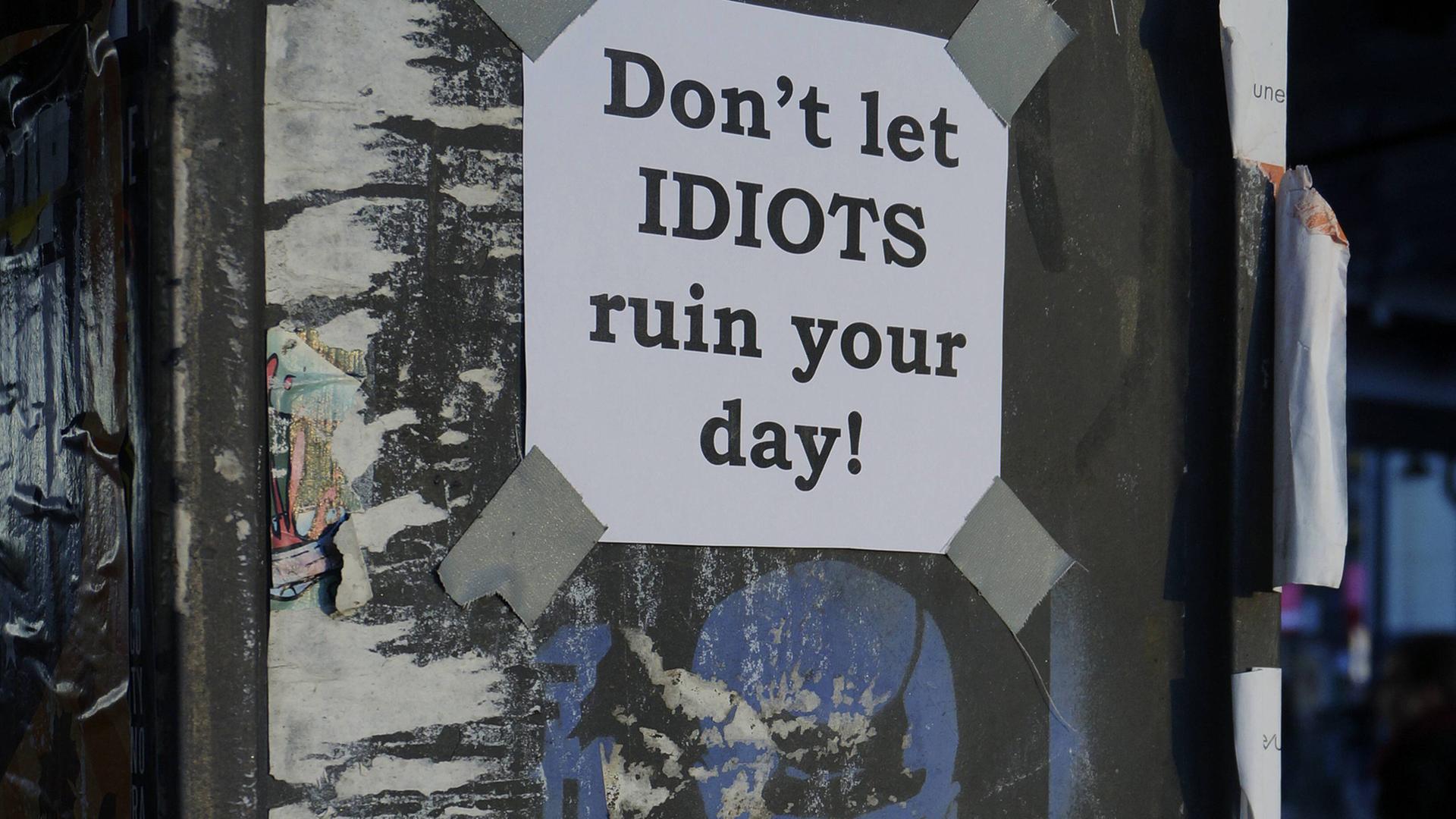 Schild mit der Aufschrift "Don´t let IDIOTS ruin your day" oberhalb eines Graffitos, das einen männlichen Kopf zeigt. Schild mit der Aufschrift "Don´t let IDIOTS ruin your day" oberhalb eines Graffitos, das einen männlichen Kopf zeigt.
