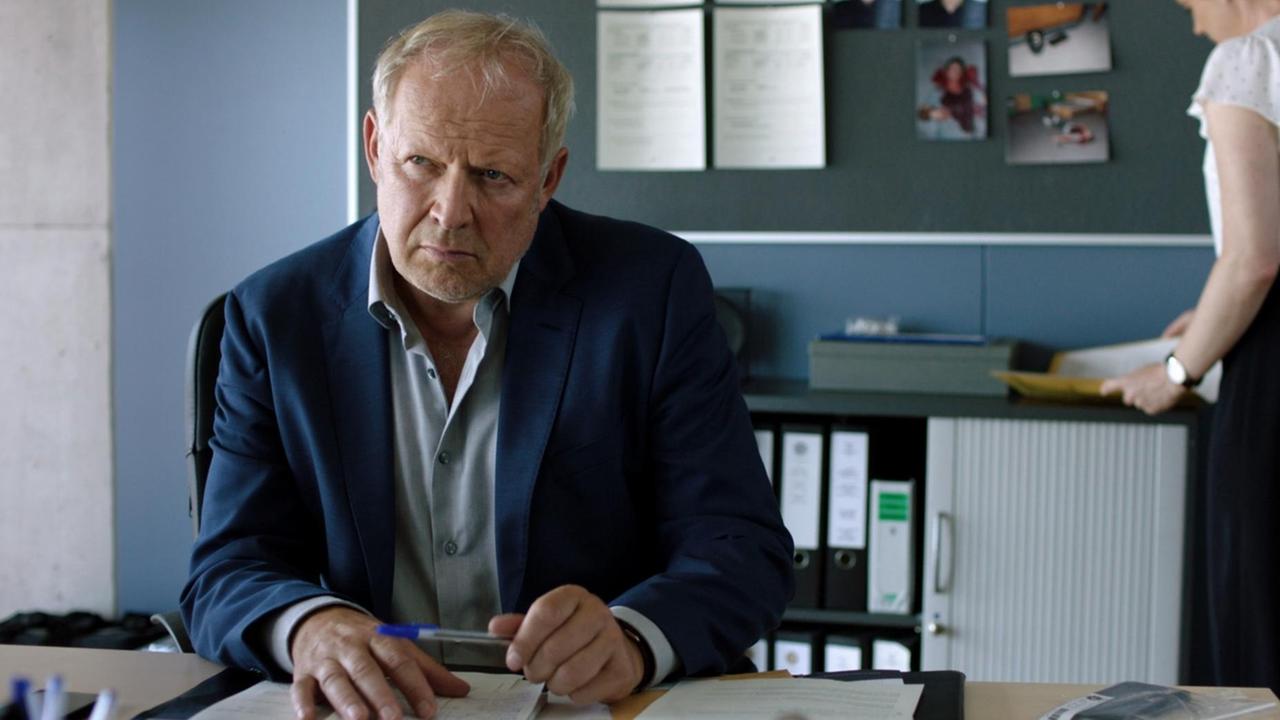 Axel Milberg sitzt im blauen Anzug an einem Tisch und blick skeptisch. Axel Milberg sitzt im blauen Anzug an einem Tisch und blick skeptisch.