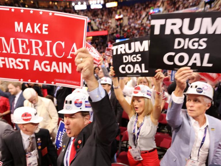 Sie sehen Frauen und Männer mit Helmen, einige halten Schilder hoch, auf einem steht "Trump gräbt nach Kohle".