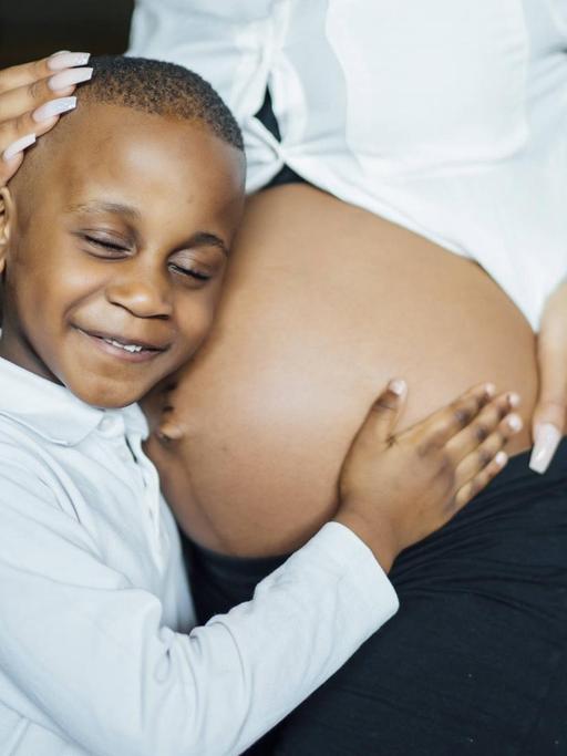 Ein kleiner Junge horcht an dem nackten Bauch einer schwangeren Frau.