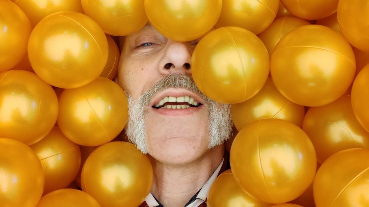 Ein Mann mit Schnauzbart ist zu erkennen, wie sein Gesicht von gelben Bällen umgegeben ist.