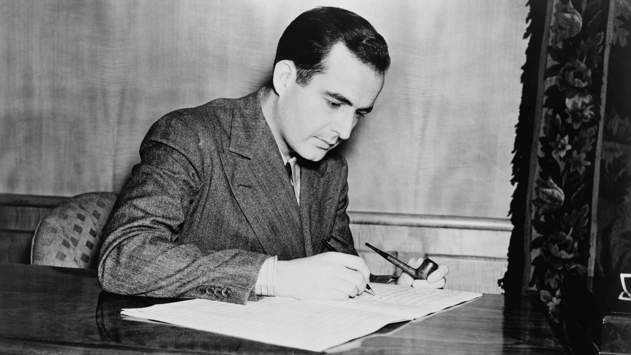 Der junge Komponist sitzt an einem Schreibtisch, er hält in der einen Hand einen Stift, mit dem er schreibt, mit der anderen eine Pfeife.