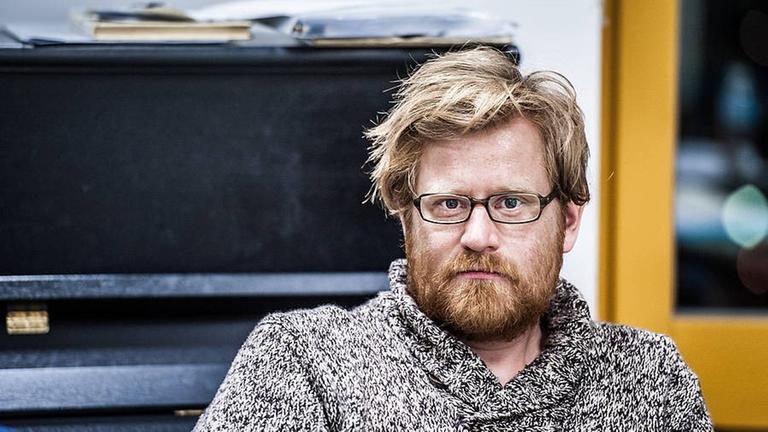 Martin Schüttler in die Kamera blickend mit Klavier im Hintergrund Martin Schüttler in die Kamera blickend mit Klavier im Hintergrund