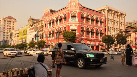 Ein Händler sitzt an einer Straße der burmesischen Metropole Yangon. Ein neue, große Autos fahren vorbei, eine Frau mit Kopftuch führt zwei Kinder an der Hand.