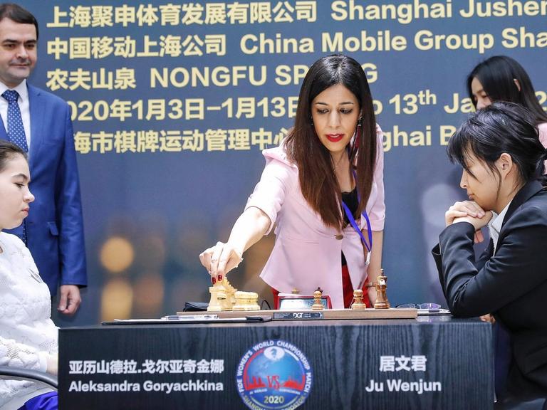 Die iranische Schiedsrichterin Shohreh Bayat bei der Schach-Weltmeisterschaft der Frauen in Schanghai. Bayat steht an einem Tisch, an dem zwei WM-Teilnehmerinnen vor einem Schachbrett sitzen, und greift nach einer Figur.