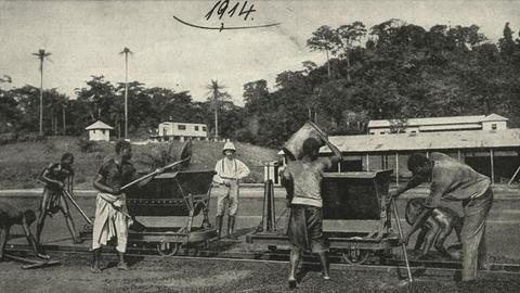 Unter Aufsicht eines deutschen Kolonisten verladen Arbeiter 1914 in Kamerun Kakaobohnen.
