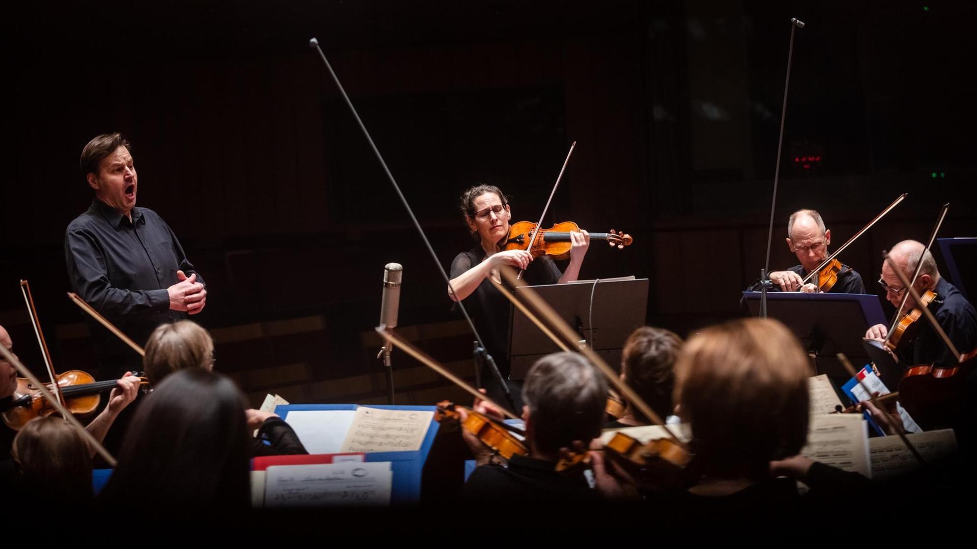 Ein Mann steht vor einem Orchester und singt. Ein Stück neben ihm steht eine Frau, spielt Violine und schaut ebenfalls in Richtung des Orchesters. Ein Mann steht vor einem Orchester und singt. Ein Stück neben ihm steht eine Frau, spielt Violine und schaut ebenfalls in Richtung des Orchesters.