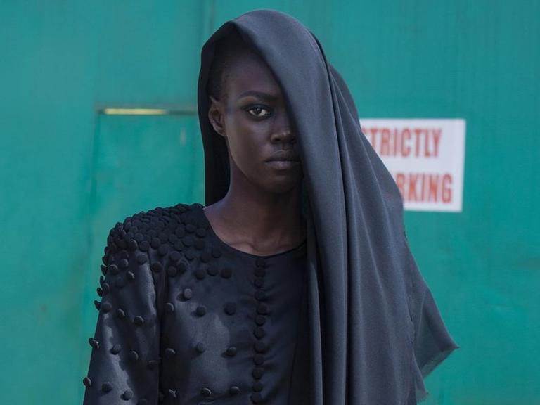Eine schwarze Frau trägt dunkle und auffällige Designer-Kleidung mit einem Kopftuch, das nur eine Seite ihres Kopfes bedeckt.