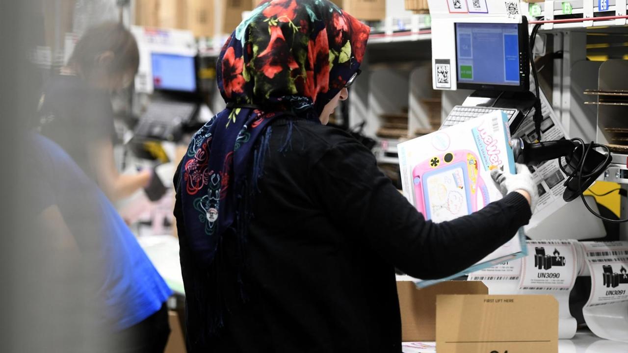 Eine Frau steht an einem Tisch und konfektioniert Pakete in einem Amazon-Verteilzentrum. Sie trägt ein Kopftuch und wird von hinten gezeigt.