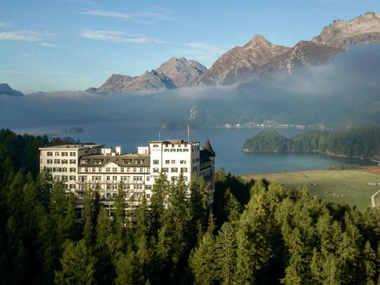 Zu sehen ist das Belle-Époque-Hotel Waldhaus Sils in einer atemberaubenden Umgebung, umrahmt von Bergen, einem See und Wäldern.