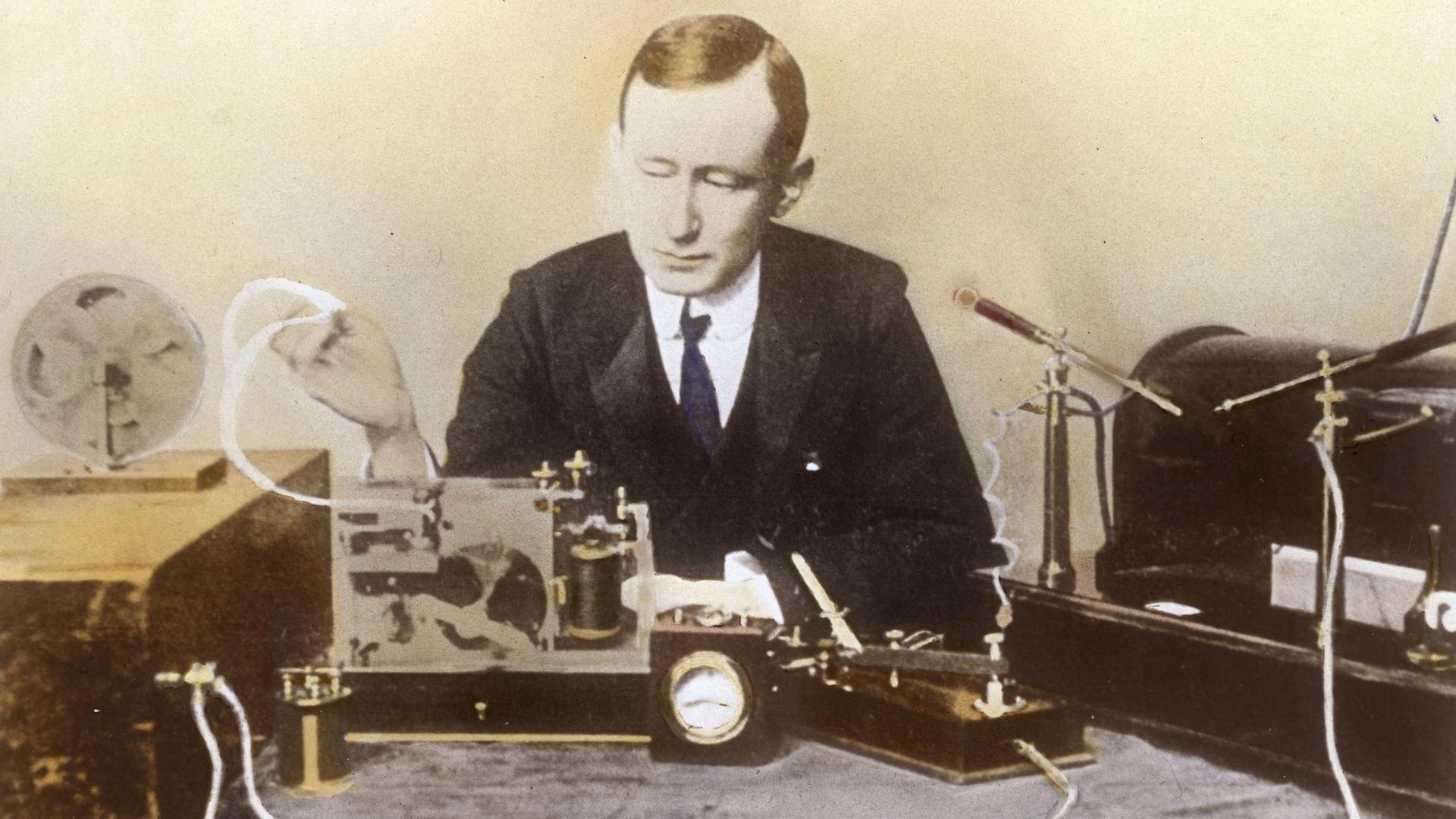 Ein nachträglich koloriertes Foto zeigt einen Mann mittleren Alters an einem Tisch vor verschiedenen Geräten wie einer Platte mit verschiedenen Drahtspulen, einer Uhr und einem historischen Mikrofon sitzend Guglielmo Marconi 1902 bei den Versuchen einer ersten transatlantischen Radio- Übertragung Ein nachträglich koloriertes Foto zeigt einen Mann mittleren Alters an einem Tisch vor verschiedenen Geräten wie einer Platte mit verschiedenen Drahtspulen, einer Uhr und einem historischen Mikrofon sitzend Guglielmo Marconi 1902 bei den Versuchen einer ersten transatlantischen Radio- Übertragung