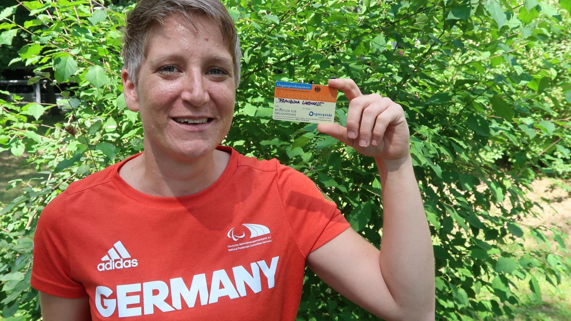 Franziska Liebhardt hält ihren Organspendeausweis in die Höhe. Sie trägt ein rotes T-Shirt mit der Aufschrift Germany. Franziska Liebhardt hält ihren Organspendeausweis in die Höhe. Sie trägt ein rotes T-Shirt mit der Aufschrift Germany.