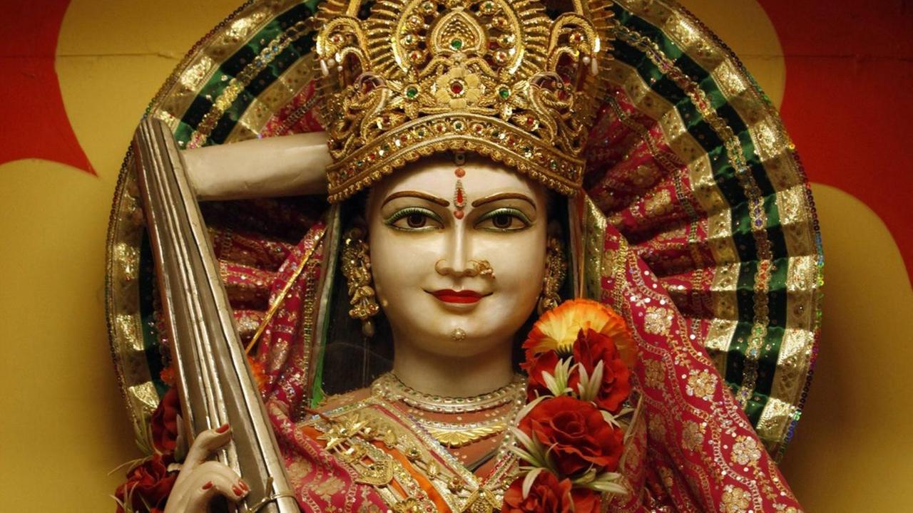 Eine Frauenfigur mit goldener Krone und einem indischen Zupfinstrument in der Hand, die mit einer Blumenranke um den Hals geschmückt wurde. Eine Frauenfigur mit goldener Krone und einem indischen Zupfinstrument in der Hand, die mit einer Blumenranke um den Hals geschmückt wurde.