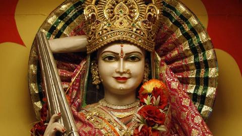 Eine Frauenfigur mit goldener Krone und einem indischen Zupfinstrument in der Hand, die mit einer Blumenranke um den Hals geschmückt wurde.