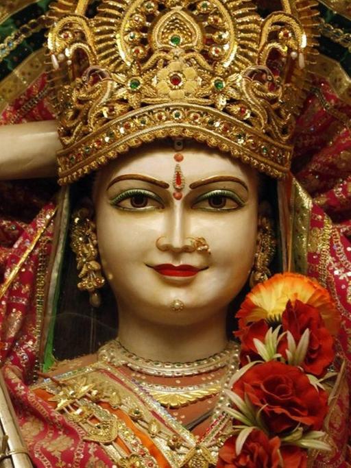 Eine Frauenfigur mit goldener Krone und einem indischen Zupfinstrument in der Hand, die mit einer Blumenranke um den Hals geschmückt wurde.