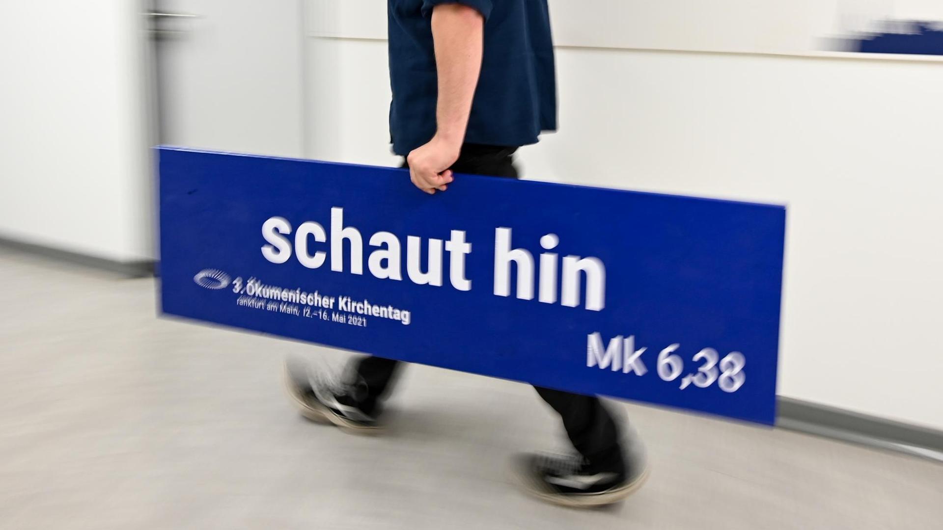Lutz Hüser, Abteilungsleiter Helfende Dienste des 3. Ökumenischen Kirchtags (ÖKT) in Frankfurt am Main, trägt in der ÖKT-Geschäftsstelle ein Schild mit der Aufschrift "schaut hin". Die Coronapandemie hat die Organisatoren des 3. Ökumenischen Kirchentags zum Umplanen gezwungen. Sie setzen nun auf vorwiegend digitale Treffen. Lutz Hüser, Abteilungsleiter Helfende Dienste des 3. Ökumenischen Kirchtags (ÖKT) in Frankfurt am Main, trägt in der ÖKT-Geschäftsstelle ein Schild mit der Aufschrift "schaut hin". Die Coronapandemie hat die Organisatoren des 3. Ökumenischen Kirchentags zum Umplanen gezwungen. Sie setzen nun auf vorwiegend digitale Treffen.